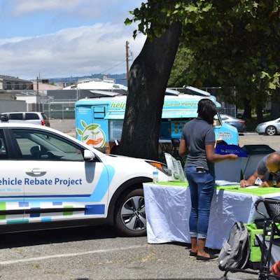 A Clean Vehicle Rebate Project event in Oakland. The image shows an electric vehicle and a man signing up for the program.