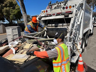 Photo of two people in high visibility vests loading debris into a packer vehicle